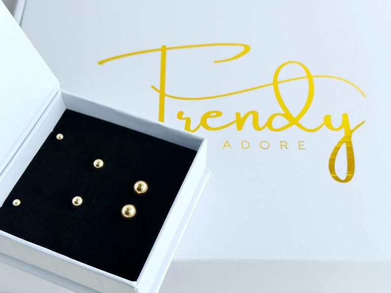 May include: A set of five gold-toned spherical stud earrings arranged on a black velvet insert inside a white square box. The box lid features the yellow text Trendy Adore. The earrings vary in size, presenting a polished, reflective metallic finish.