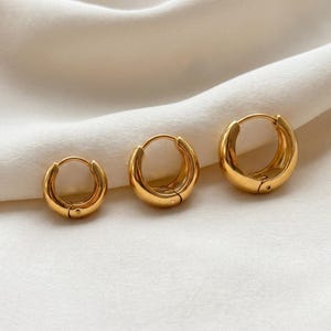 3 Gold-colored hoop earrings, 8mm, 10mm 12mm inner diameter size with their outer diameter being 15, 16, and 18mm respectively, are displayed upright on a soft ivory cream fabric. The image is a close-up, highlighting the jewelry's design and shine.