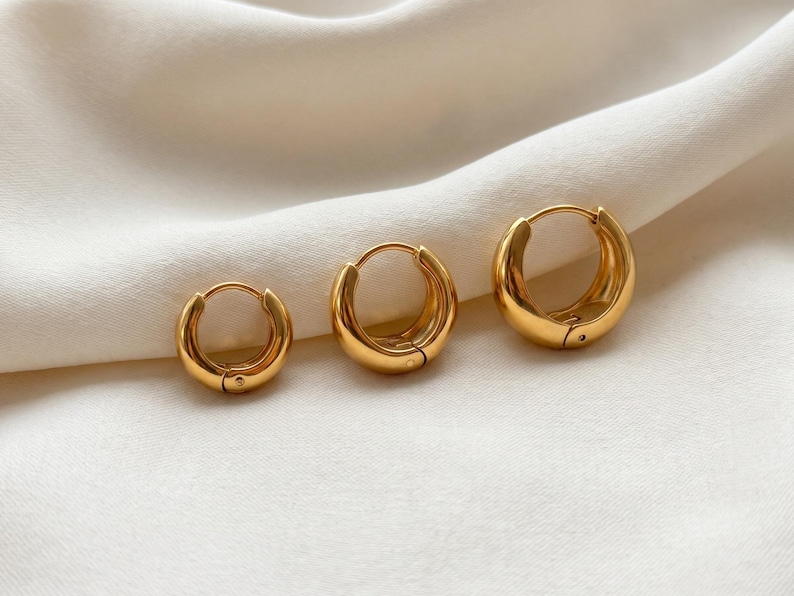 3 Gold-colored hoop earrings, 8mm, 10mm 12mm inner diameter size with their outer diameter being 15, 16, and 18mm respectively, are displayed upright on a soft ivory cream fabric. The image is a close-up, highlighting the jewelry's design and shine.