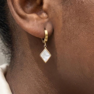 May include: A gold-colored hoop earring with a diamond-shaped pendant. The pendant has a white, iridescent center. The earring is shown on a person's ear.