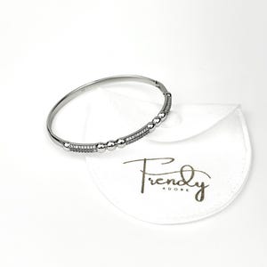 May include: A silver bangle bracelet with a row of small, clear stones and a row of larger, clear stones. The bracelet is on a white background with a white pouch that says "Trendy Adore".