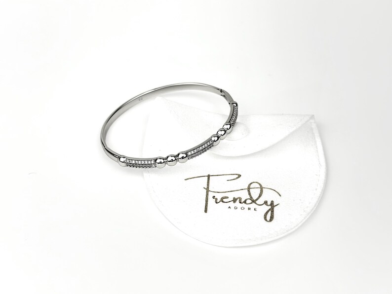 May include: A silver bangle bracelet with a row of small, clear stones and a row of larger, clear stones. The bracelet is on a white background with a white pouch that says "Trendy Adore".