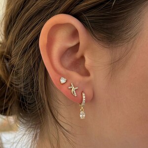 May include: Close-up of an ear adorned with three gold earrings. The top earring is a heart-shaped stud. The middle earring is a cross design. The bottom earring is a small hoop with a dangling teardrop-shaped charm, all encrusted with small, clear stones.