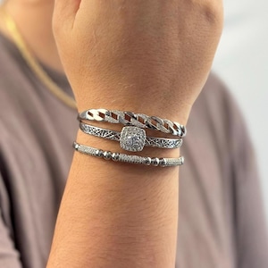 May include: A set of three silver bracelets with a square-shaped, clear crystal centerpiece. The bracelets are stacked on a person's wrist.