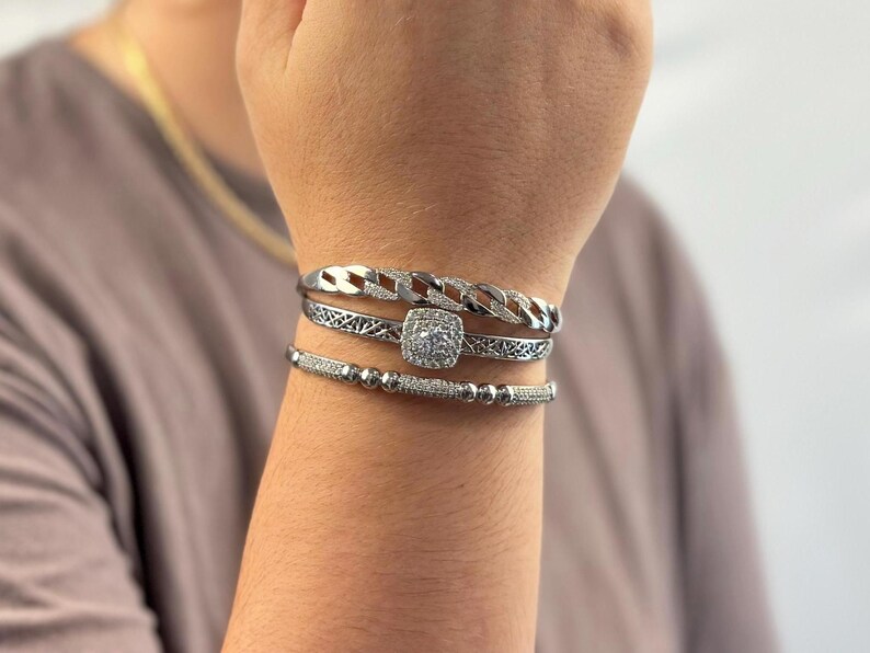 May include: A set of three silver bracelets with a square-shaped, clear crystal centerpiece. The bracelets are stacked on a person's wrist.