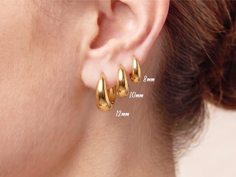 Gold-colored hoop earrings, 8mm, 10mm and 12mm in size, are displayed on an ear. The earrings have a smooth, polished finish and a classic, rounded shape. The image is a close-up, highlighting the jewelry's design and shine.