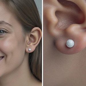 May include: Close-up of a woman's ear with a small, round, silver stud earring. The earring has a textured, glittery surface. The woman is smiling, and her other ear also has a matching earring.