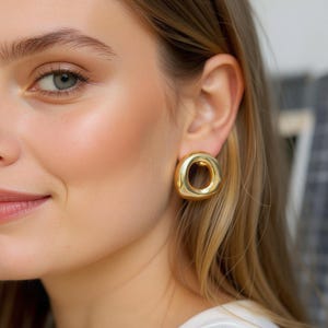 May include: Gold-toned, circular stud earrings. The earrings have a smooth, polished finish and a hollow center. The earrings are worn on a person's ear. The background is blurred.