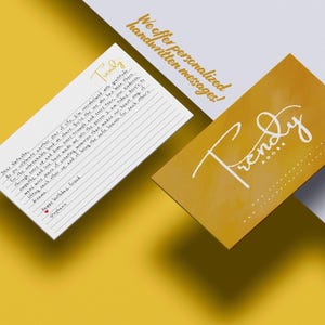 May include: A white rectangular card with lined paper and handwritten text, alongside a mustard yellow business card featuring the white script logo Trendy Adore. The text on the card includes the word Trendy and a message ending with Happy birthday, friend.
