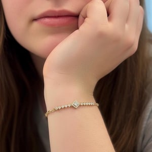 May include: A gold bracelet with a diamond-shaped centerpiece and small, round accent stones. The bracelet is worn on a wrist, showcasing its delicate design and sparkle. The jewelry piece is a stylish accessory.