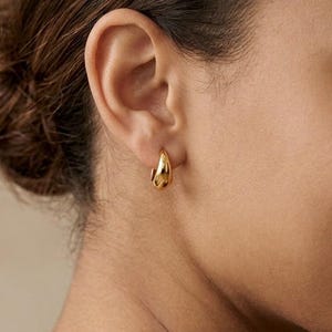 Gold-colored hoop earrings, 10mm in size, are displayed on an ear. The earrings have a smooth, polished finish and a classic, rounded shape. The image is a close-up, highlighting the jewelry's design and shine.