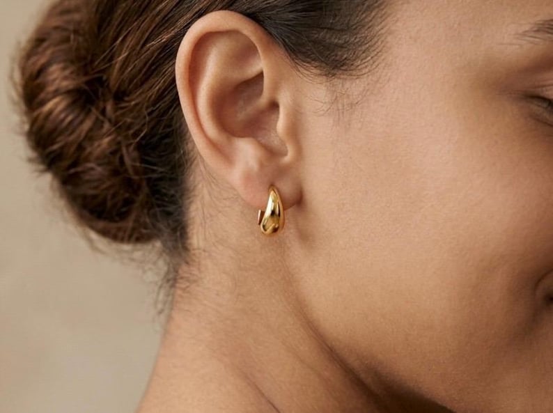 Gold-colored hoop earrings, 10mm in size, are displayed on an ear. The earrings have a smooth, polished finish and a classic, rounded shape. The image is a close-up, highlighting the jewelry's design and shine.