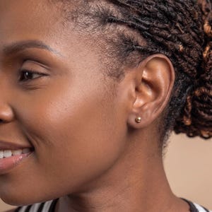May include: A small, polished gold ball stud earring worn in the earlobe. The earring features a simple, spherical design with a smooth, reflective metallic surface that catches the light, providing a subtle and classic appearance for daily wear.