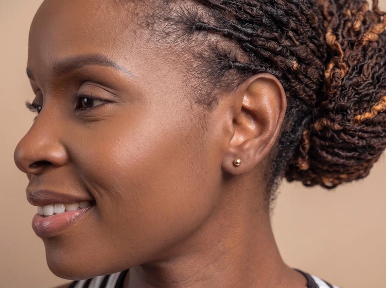May include: A small, polished gold ball stud earring worn in the earlobe. The earring features a simple, spherical design with a smooth, reflective metallic surface that catches the light, providing a subtle and classic appearance for daily wear.