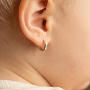 May include: Close-up of a child's ear with a small gold hoop earring. The earring is adorned with several small, clear gemstones. The child's skin is fair, and the earring is the focal point of the image.