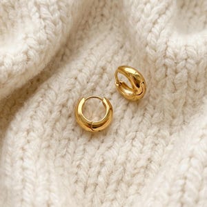 Gold-colored hoop earrings, 8mm in size, are displayed on a hand woven ivory cream fabric. The earrings have a smooth, polished finish and a classic, rounded shape. The image is a close-up, highlighting the jewelry's design and shine.