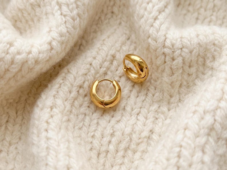 Gold-colored hoop earrings, 8mm in size, are displayed on a hand woven ivory cream fabric. The earrings have a smooth, polished finish and a classic, rounded shape. The image is a close-up, highlighting the jewelry's design and shine.