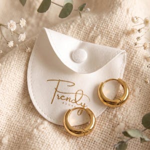 Gold-colored hoop earrings, 12mm in size, are displayed next to trendy adore jewelry pouch on a hand woven ivory cream fabric. The earrings have a smooth, polished finish and a classic, rounded shape. The image is a close-up, highlighting the jewelry's design and shine.