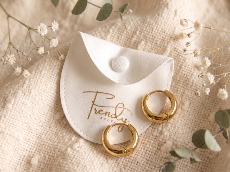 Gold-colored hoop earrings, 12mm in size, are displayed next to trendy adore jewelry pouch on a hand woven ivory cream fabric. The earrings have a smooth, polished finish and a classic, rounded shape. The image is a close-up, highlighting the jewelry's design and shine.