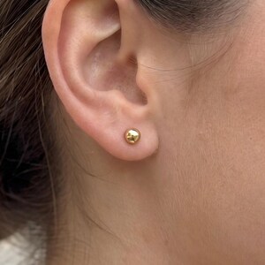 May include: A small, polished gold ball stud earring worn in the earlobe. The earring features a smooth, spherical design with a reflective metallic finish, providing a simple and classic appearance that sits flush against the skin of the ear.