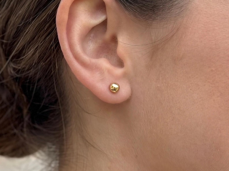May include: A small, polished gold ball stud earring worn in the earlobe. The earring features a smooth, spherical design with a reflective metallic finish, providing a simple and classic appearance that sits flush against the skin of the ear.