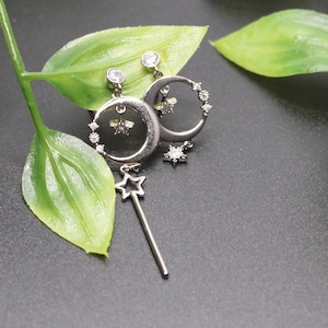May include: Silver-toned earrings featuring a crescent moon design with embedded clear crystals and a dangling star-shaped charm. The earrings have a post-back closure and are set against a dark background with green leaves.