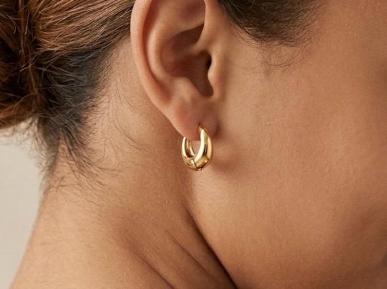 Gold-colored hoop earrings, 12mm in size, are displayed on an ear. The earrings have a smooth, polished finish and a classic, rounded shape. The image is a close-up, highlighting the jewelry's design and shine.