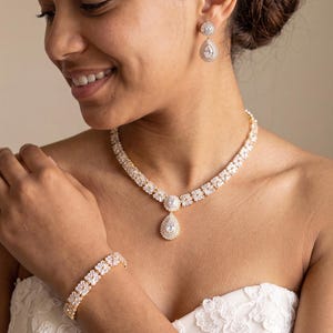 May include: A jewellery set consisting of a necklace, a bracelet, and drop earrings. Each piece features clear, faceted stones set in a gold-toned metal frame. The necklace includes a central teardrop-shaped pendant, matching the design of the earrings.
