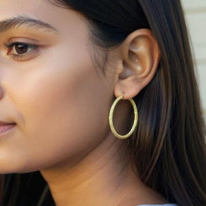 Pode incluir: Brincos de argola ovais dourados com superfície texturizada. Os brincos são usados na orelha de uma pessoa com cabelo escuro e tez clara. Design simples e elegante.