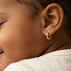 Puede incluir: Primer plano de la oreja de un niño con un pendiente de aro dorado. El pendiente tiene un diseño pequeño y redondeado, y el color dorado es brillante y brillante. La piel del niño es de un tono marrón cálido, y el pendiente es el punto focal de la imagen.
