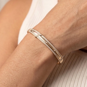 May include: A gold-toned hinged bangle bracelet featuring a textured center panel with a repeating pattern, bordered by rows of small clear stones. The bracelet has a polished finish and a secure clasp mechanism visible on the top side of the band.