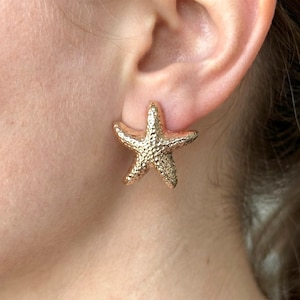 May include: Gold-colored starfish stud earrings. The earrings are textured and have a detailed, realistic design. The earrings are worn on an ear, showcasing their size and shape. The earrings are a beach-themed accessory.
