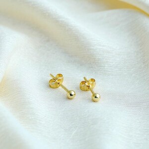 May include: A pair of gold stud earrings featuring a small, polished spherical ball at the end of a straight post, accompanied by matching butterfly-style earring backs. The earrings have a bright, reflective metallic finish and a simple design.