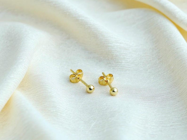 May include: A pair of gold stud earrings featuring a small, polished spherical ball at the end of a straight post, accompanied by matching butterfly-style earring backs. The earrings have a bright, reflective metallic finish and a simple design.