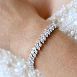May include: A silver bracelet set with numerous clear, teardrop-shaped gemstones. The bracelet, worn on a wrist, features a design resembling connected leaves. The background shows a white, beaded garment.
