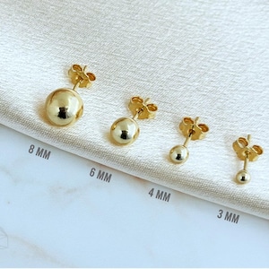 May include: Four gold-tone ball stud earrings arranged in a row, labeled with sizes 8 mm, 6 mm, 4 mm, and 3 mm. Each earring features a smooth, spherical design with a post back, showcasing a polished metallic finish and varying diameters.