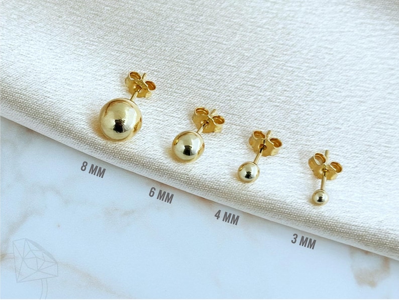 May include: Four gold-tone ball stud earrings arranged in a row, labeled with sizes 8 mm, 6 mm, 4 mm, and 3 mm. Each earring features a smooth, spherical design with a post back, showcasing a polished metallic finish and varying diameters.