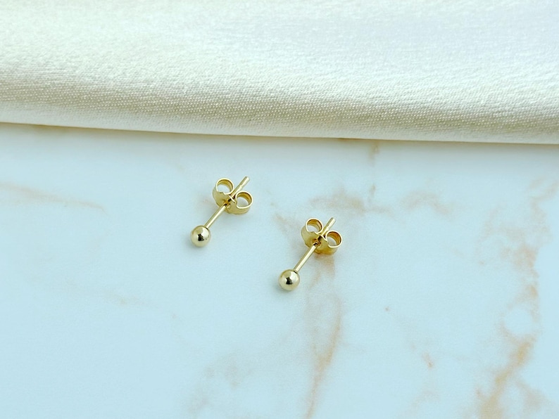 May include: A pair of gold ball stud earrings featuring a small, polished spherical bead attached to a thin post with a matching butterfly clutch backing. The earrings have a bright, reflective metallic finish and a simple, classic design.