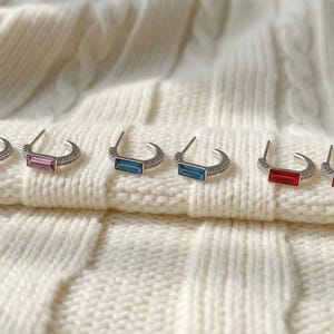 May include: Six silver-toned hoop earrings with rectangular gemstones in pink, blue, and red. The earrings are set on a white knitted surface, showcasing their design and color variations. Each earring has a small, clear stone accent.
