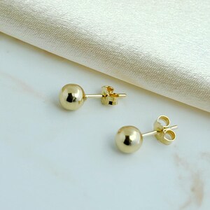 May include: A pair of polished gold ball stud earrings with matching butterfly backings. Each earring features a smooth, spherical design with a reflective metallic finish, showcasing a simple and classic circular shape for the earlobe.