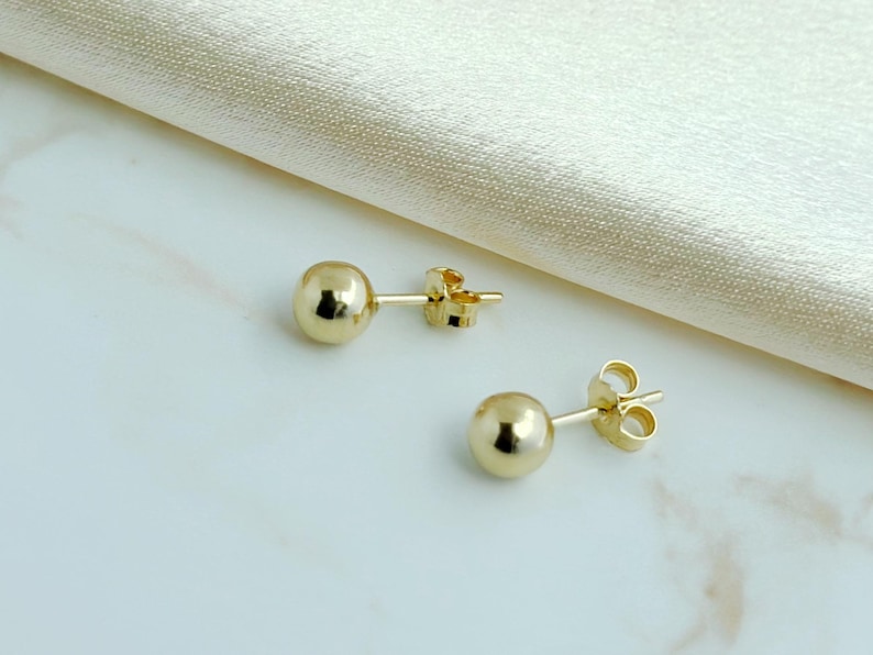 May include: A pair of polished gold ball stud earrings with matching butterfly backings. Each earring features a smooth, spherical design with a reflective metallic finish, showcasing a simple and classic circular shape for the earlobe.