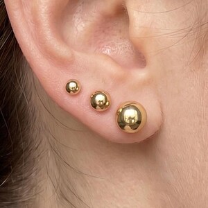 May include: Three polished gold ball stud earrings of varying sizes arranged in a row along the earlobe. Each earring features a smooth, spherical design with a reflective metallic finish, creating a graduated look from the smallest to largest.