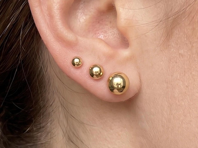 May include: Three polished gold ball stud earrings of varying sizes arranged in a row along the earlobe. Each earring features a smooth, spherical design with a reflective metallic finish, creating a graduated look from the smallest to largest.