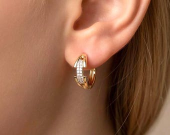 May include: A close-up of a gold hoop earring with a row of small, sparkling white stones set in the center. The earring is worn on a person's ear, with the skin visible in the background. The earring has a modern, elegant design.