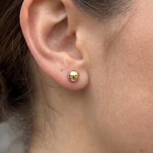 May include: A small, polished gold-tone ball stud earring worn in the earlobe. The earring features a smooth, spherical design with a reflective metallic surface that catches the light, creating a simple and classic circular shape on the skin.