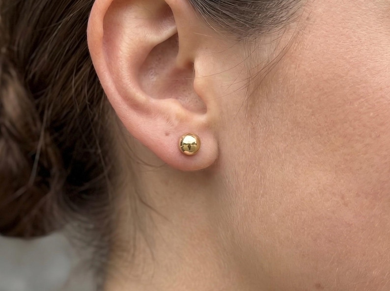 May include: A small, polished gold-tone ball stud earring worn in the earlobe. The earring features a smooth, spherical design with a reflective metallic surface that catches the light, creating a simple and classic circular shape on the skin.