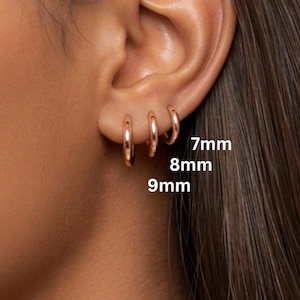 May include: Close-up of three rose gold hoop earrings, 7mm, 8mm, and 9mm in size, displayed on an ear. The earrings are simple, circular, and shiny. The background is a blurred view of skin and hair.