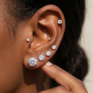 May include: Close-up of an ear adorned with multiple diamond stud earrings of varying sizes. The earrings are set in a classic, minimalist style, reflecting light and adding sparkle. The ear is set against a neutral background.