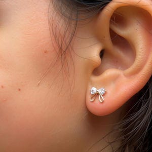 May include: A close-up of a person's ear with a small, gold-colored bow-shaped stud earring. The earring features two clear, round gemstones. The skin around the ear is light-toned.