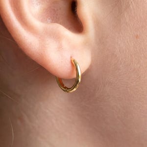 Peut inclure: Gros plan d'une boucle d'oreille créole dorée portée à l'oreille. La boucle d'oreille est de conception simple et circulaire, avec une surface lisse et polie. La boucle d'oreille est de couleur dorée chaude et semble être en métal. La boucle d'oreille est fixée par un fermoir à charnière.